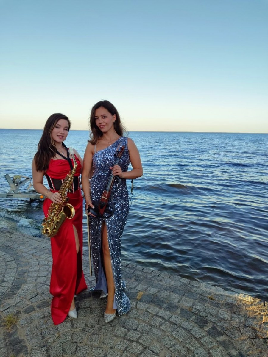 Iva Sax в красивом красном концертном платье играет на золотистом саксофоне на живописной площадке среди хвойных деревьев возле Киевского моря