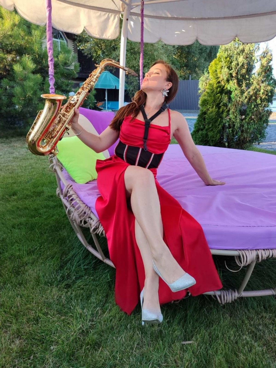 Саксофоністка Iva Sax виступає на відкритому майданчику біля Київського моря серед хвойного саду у розкішній червоній концертній сукні із золотистим саксофоном.